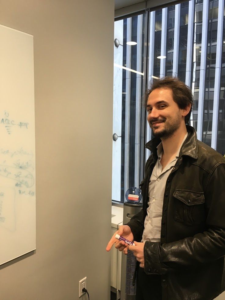 A person in a leather jacket holding a marker and smiling, standing in front of a whiteboard in an office with large windows.