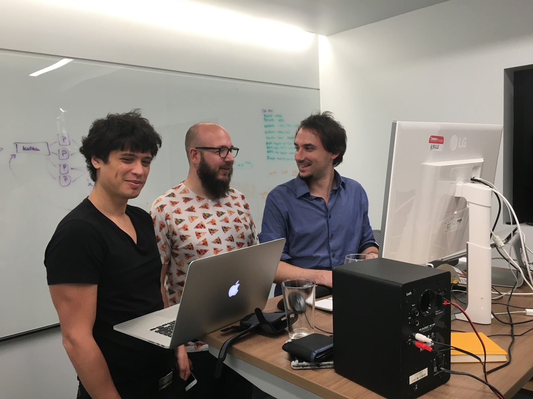 Three people standing and smiling in front of computers in an office, with a whiteboard behind them.