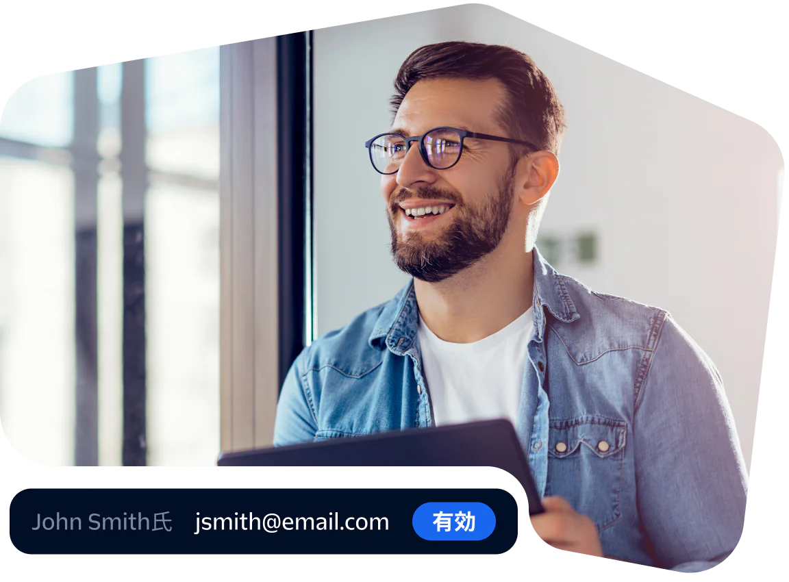 Man in glasses and denim shirt smiling while using a tablet near a window.