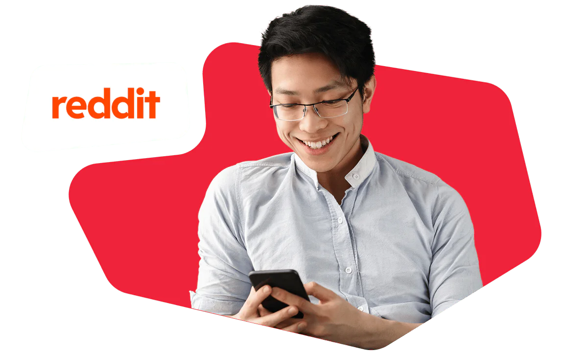 A man with glasses smiles while looking at a smartphone, with a Reddit logo in the background.