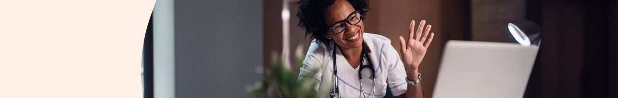 Healthcare professional with stethoscope smiling and waving at laptop during a video call.