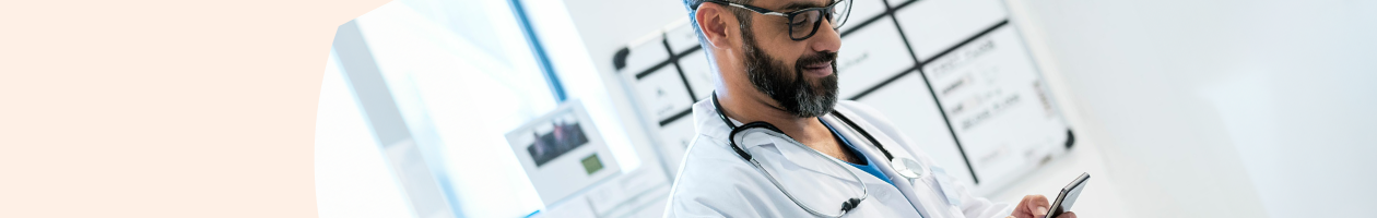 Male doctor wearing glasses and stethoscope looks at his cellphone in a hospital setting.