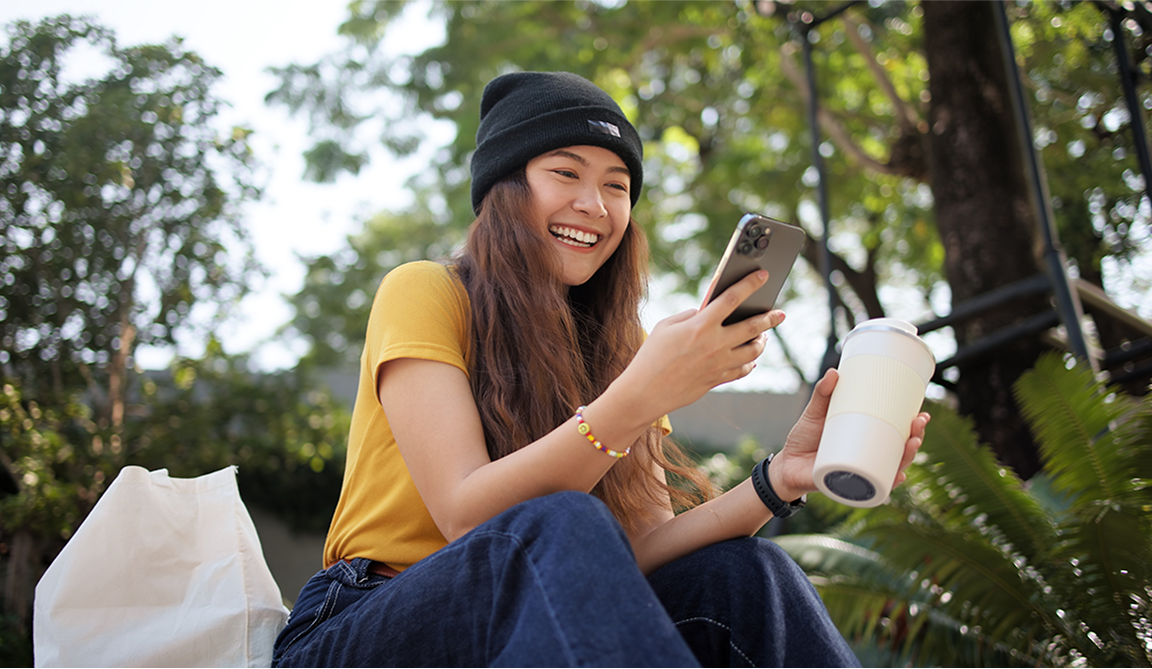 Woman with long brown hair wearing a beanie and yellow shirt, holding a phone and coffee cup, smiling outdoors.