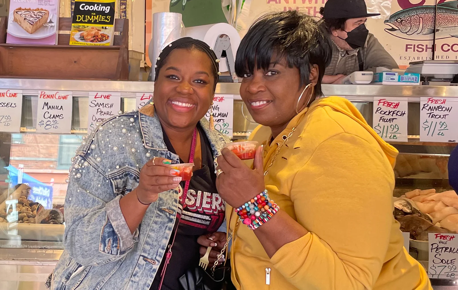 Two smiling women holding clam chowder shots inside a fish market.
