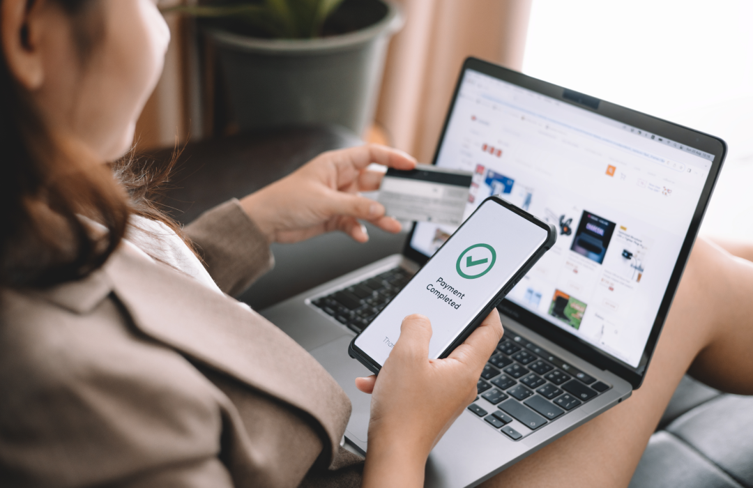 Woman confirms payment on smartphone while shopping online on a laptop.