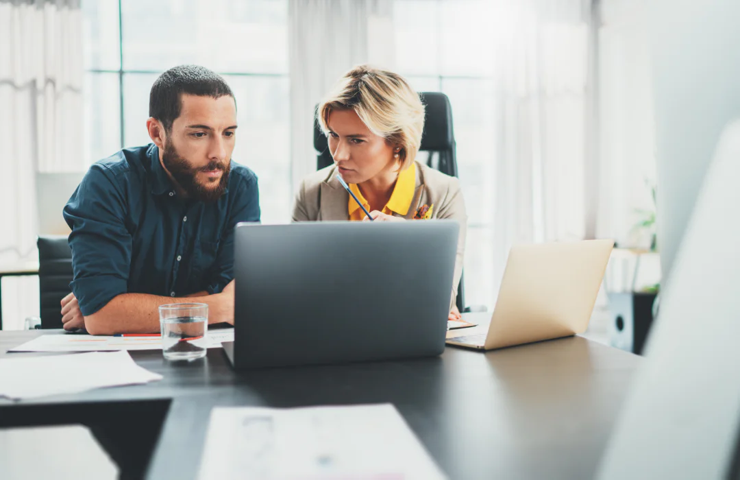 Two colleagues working together at a desk with laptops in a bright office.