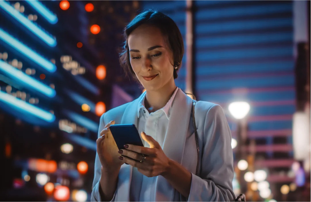 Woman looks at smartphone while standing on city street at night.