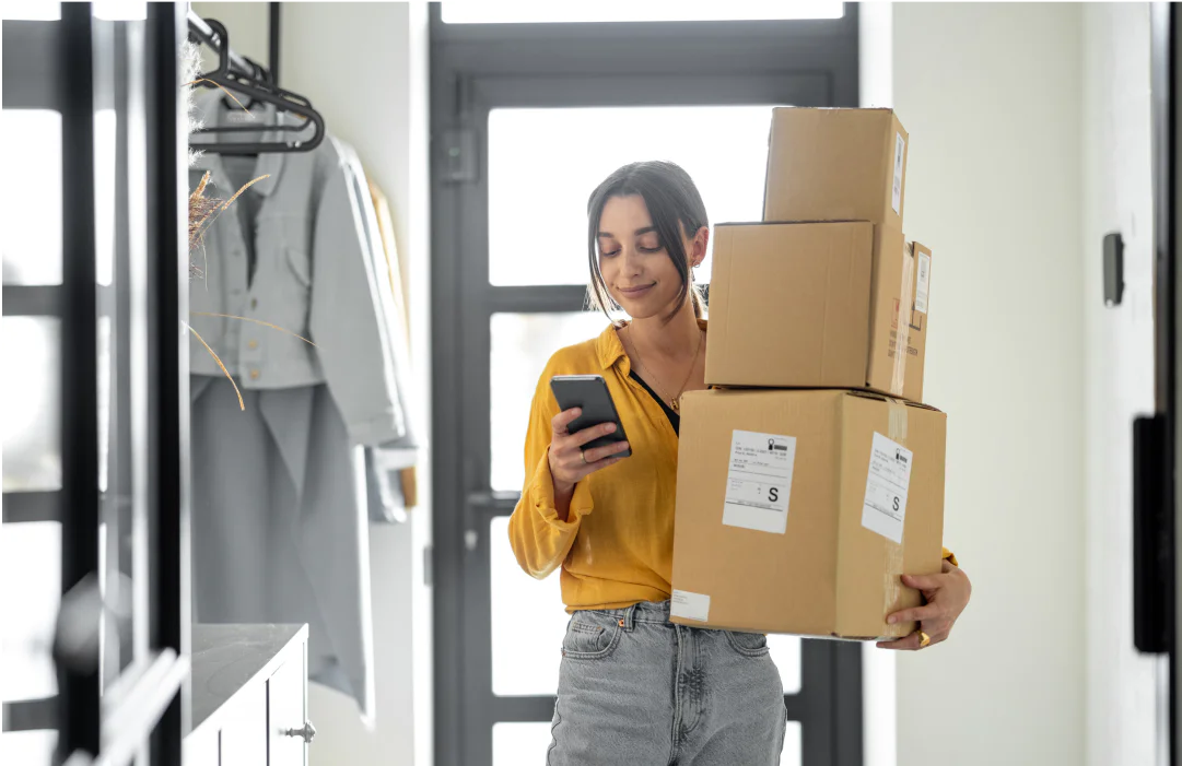 A woman in a yellow shirt and jeans carries three boxes and checks her phone in a bright hallway.