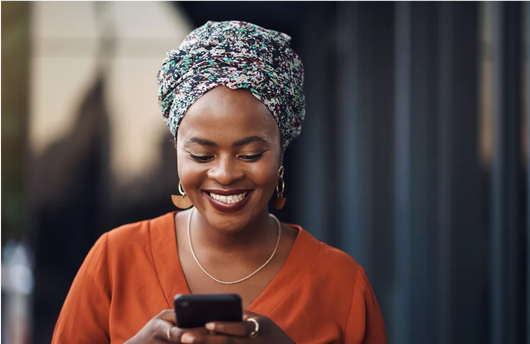 A woman with a floral head wrap and orange top smiling at her smartphone.