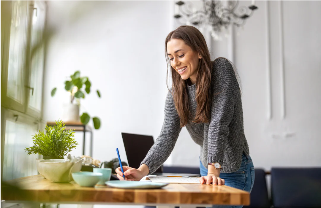 Woman writing in a notebook at a wooden desk with a laptop and plants in a well-lit room