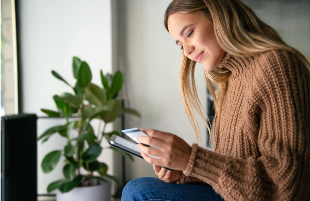 Woman holding a credit card and tablet while smiling, sitting indoors beside a potted plant.
