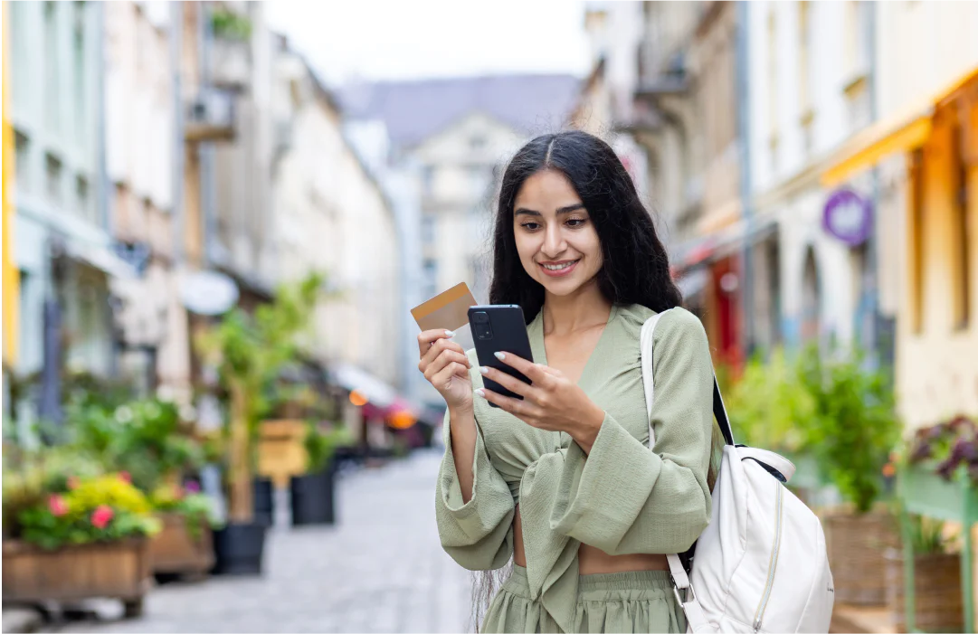 Young woman holding a smartphone and credit card, smiling, on a city street with shops.