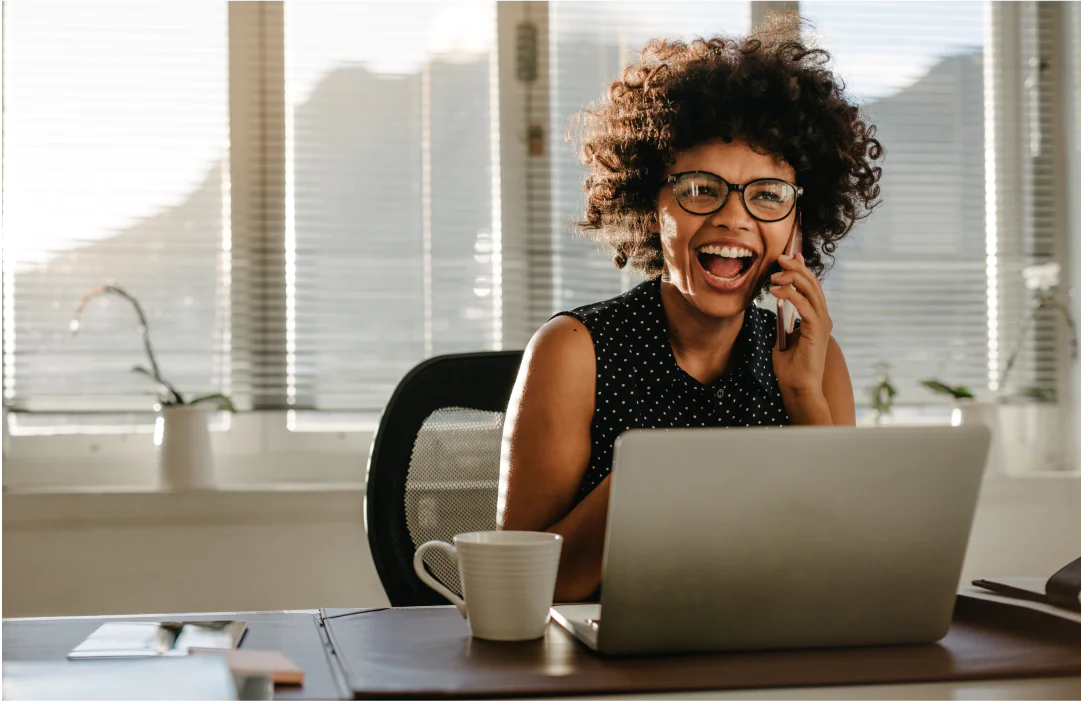 Smiling woman with curly hair on the phone, working on a laptop in a sunny office