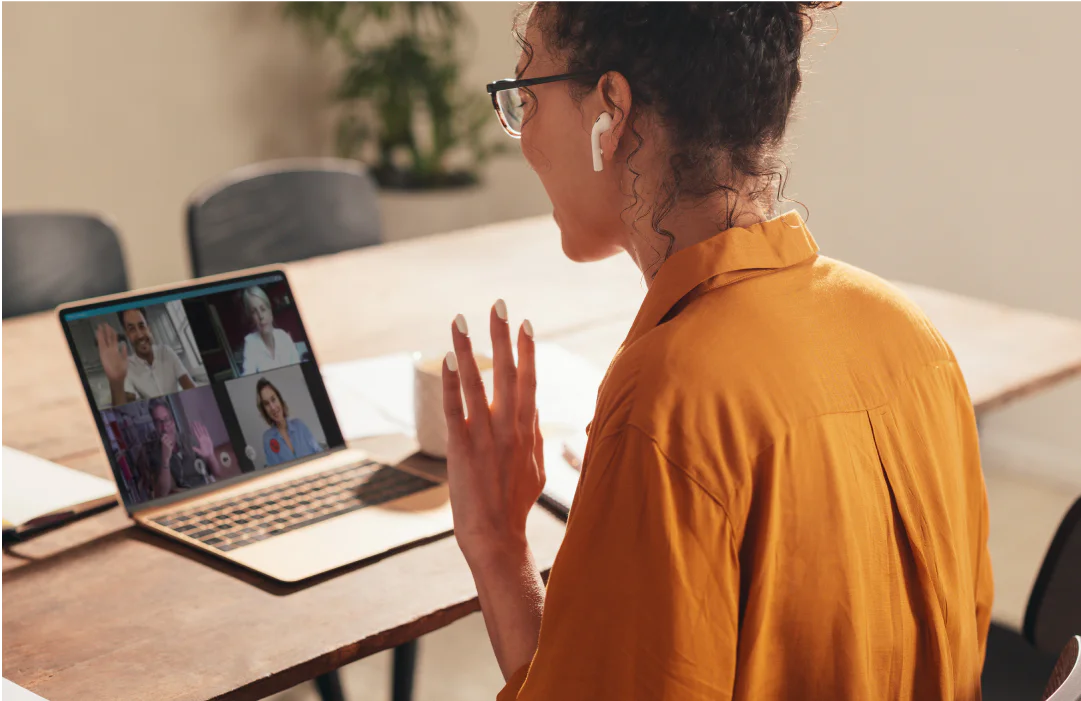 Woman wearing earbuds waving at a laptop screen with a virtual meeting of four people in progress.