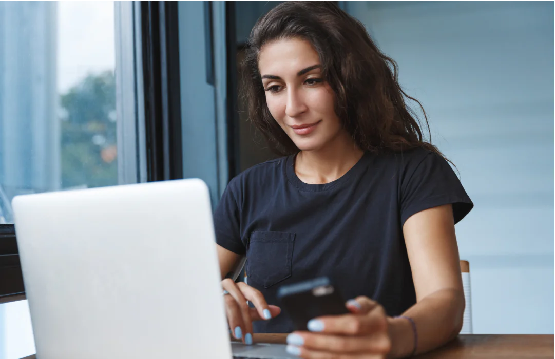 Woman using a laptop and holding a smartphone while sitting at a desk.