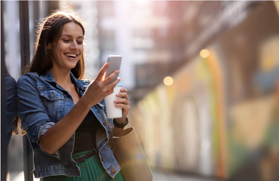 Woman in a denim jacket smiling at her smartphone while holding a cup, standing against a wall