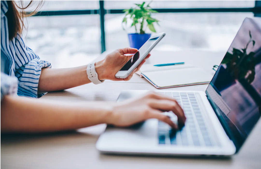 Person using a laptop and holding a smartphone at a desk with a notebook and plant nearby
