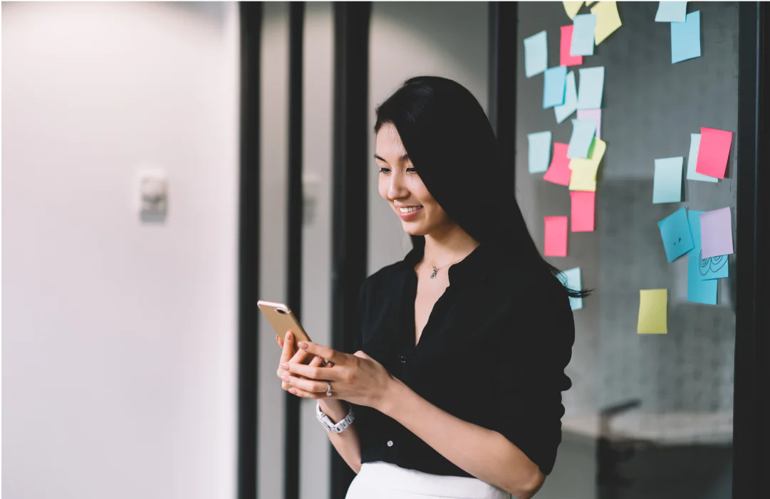 Woman in black shirt using a smartphone and smiling, standing near a wall with colorful sticky notes.