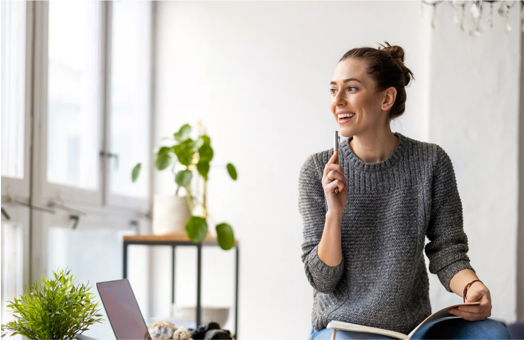 Woman sitting by a window, smiling and holding a pen, with a notebook on her lap and a laptop on the table.