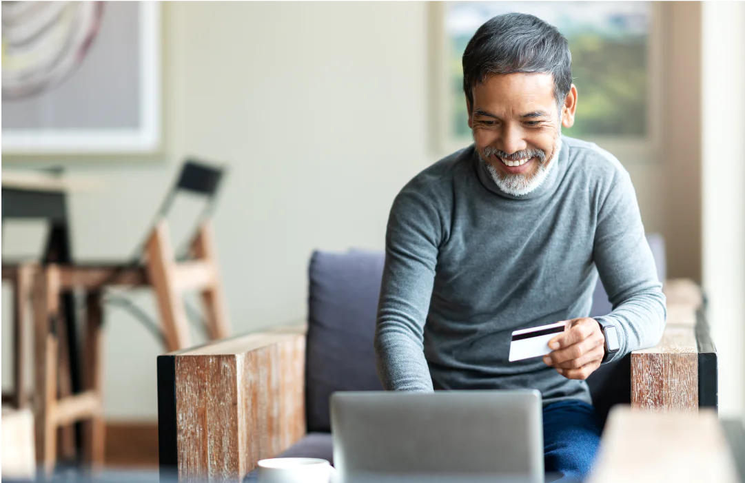 Older man smiling while holding a credit card and using a laptop in a cozy living room.