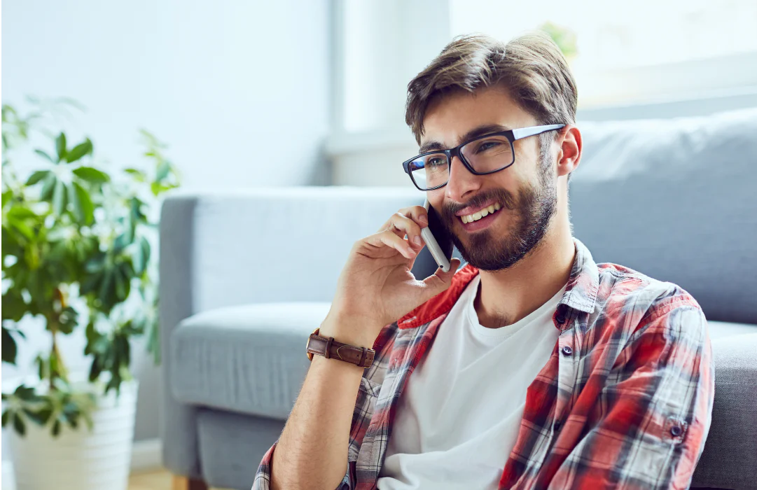 Young man with glasses and beard talking on a phone, sitting on a couch, with a green plant nearby.
