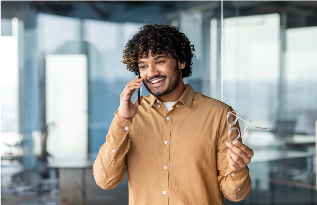 Man in a brown shirt smiling while talking on the phone and holding eyeglasses in an office setting.