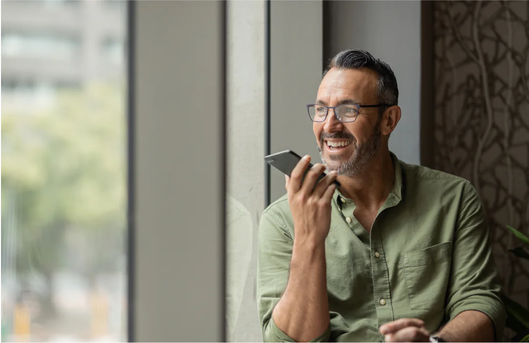 Man with glasses and a beard smiling, holding a smartphone, sitting by a window.