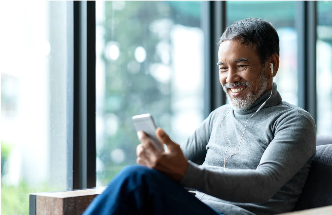 Man with earbuds smiling at smartphone near large window