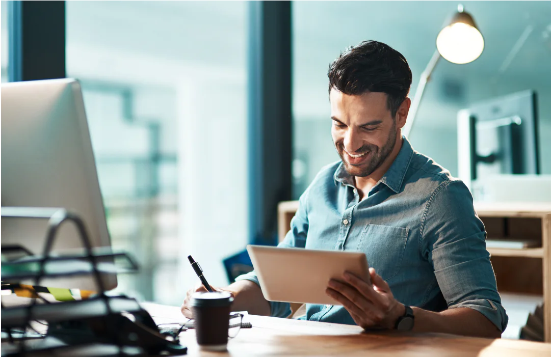 Man in blue shirt smiles while using a tablet, holding a pen, at a desk with a lamp and a coffee cup.