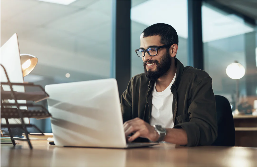 Man with glasses and beard working on a laptop at a desk in a modern office environment.