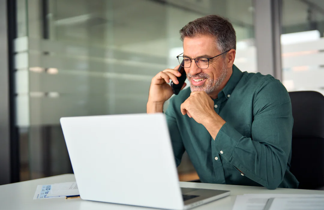 Man in glasses using a laptop and talking on a phone in a modern office setting