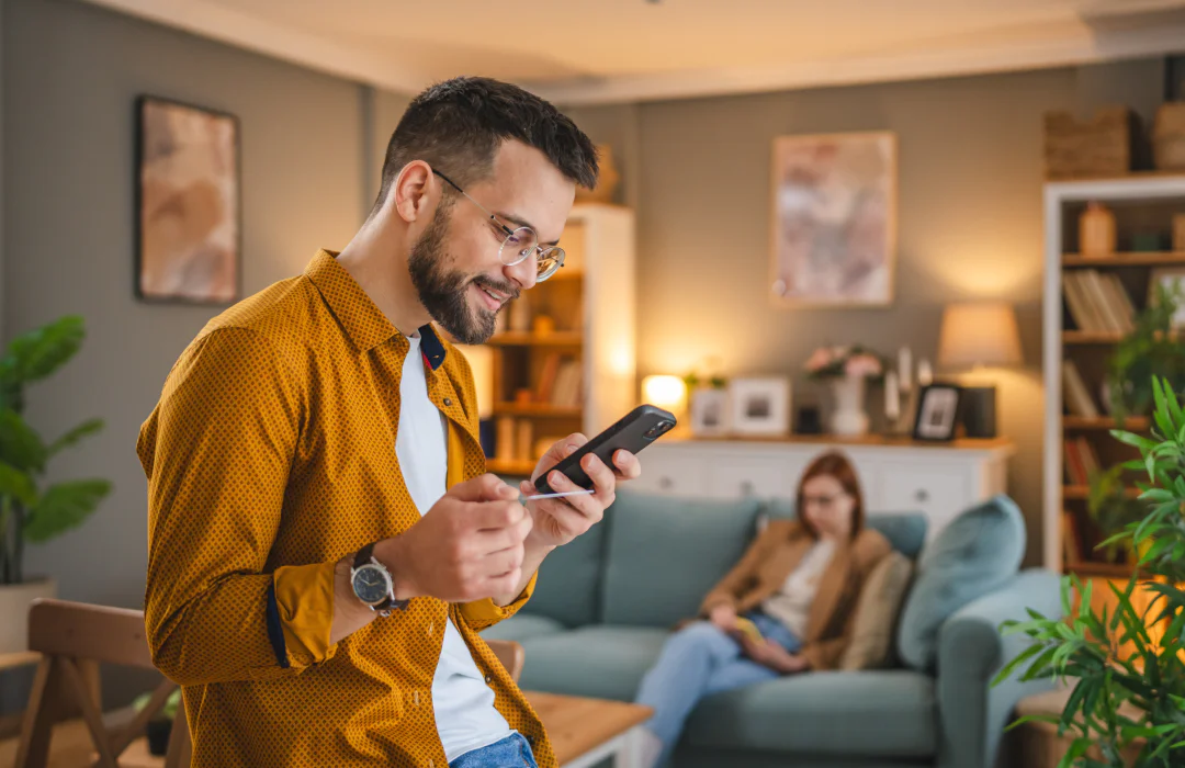 A man in glasses and a yellow shirt smiles at his smartphone in a cozy living room, with a woman on the couch.