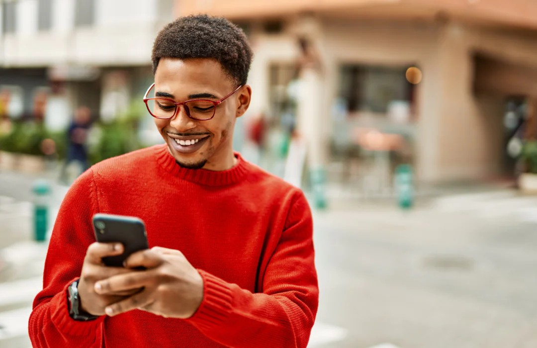 A man in a red sweater and glasses is smiling while looking at his smartphone on a city street.