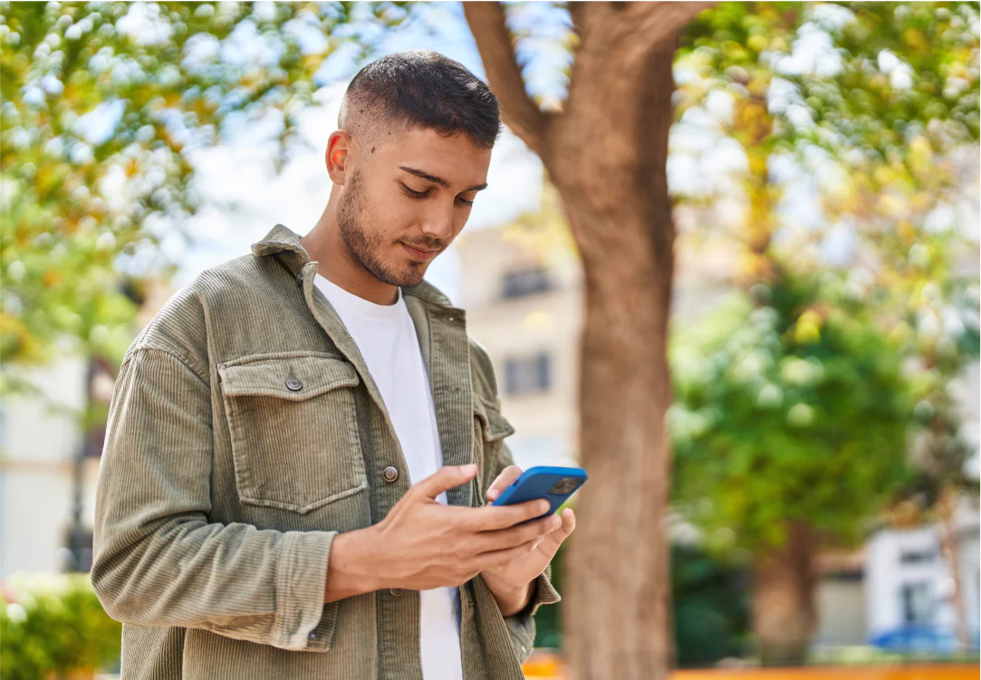 A young man in a green jacket looking at his smartphone while standing in a park.
