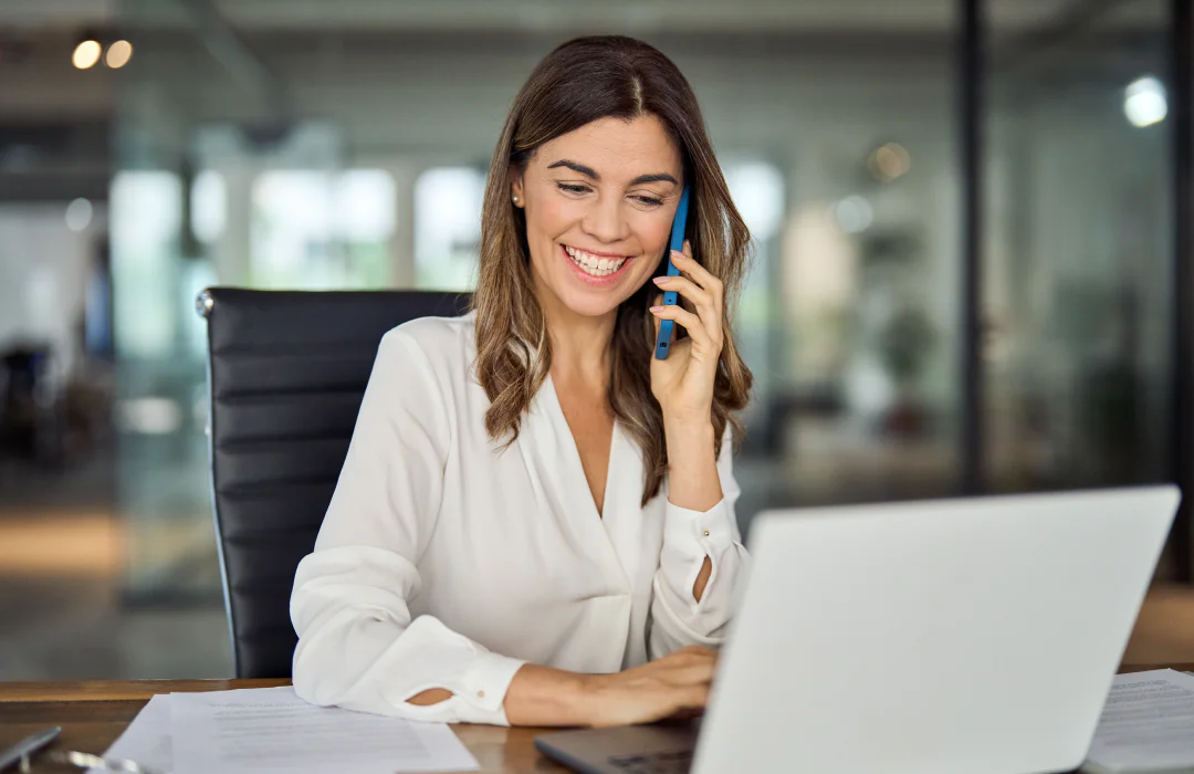Businesswoman in white blouse smiling while talking on phone and working on laptop at office desk.