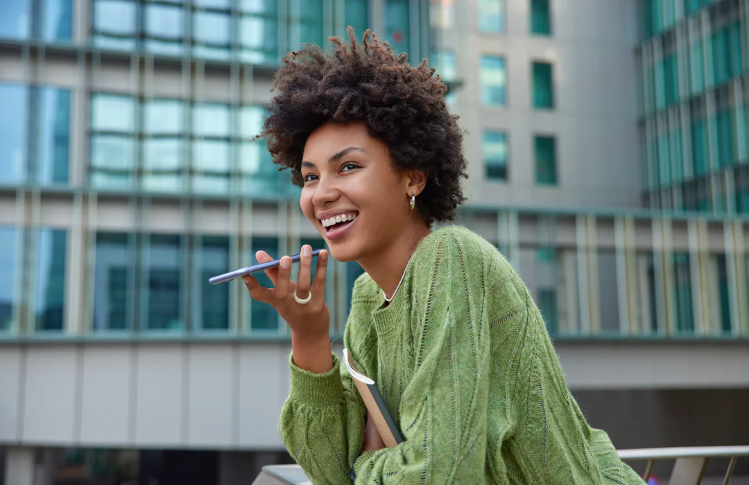 Smiling woman talking on a smartphone, standing outdoors in front of a modern building.