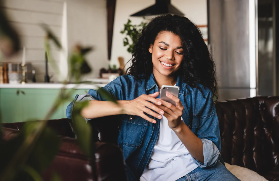 Smiling woman using a smartphone on a brown leather couch in a modern living room.