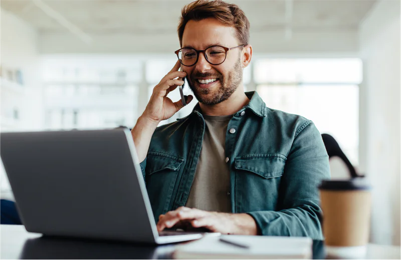 Man wearing glasses talking on phone and using laptop in a modern office