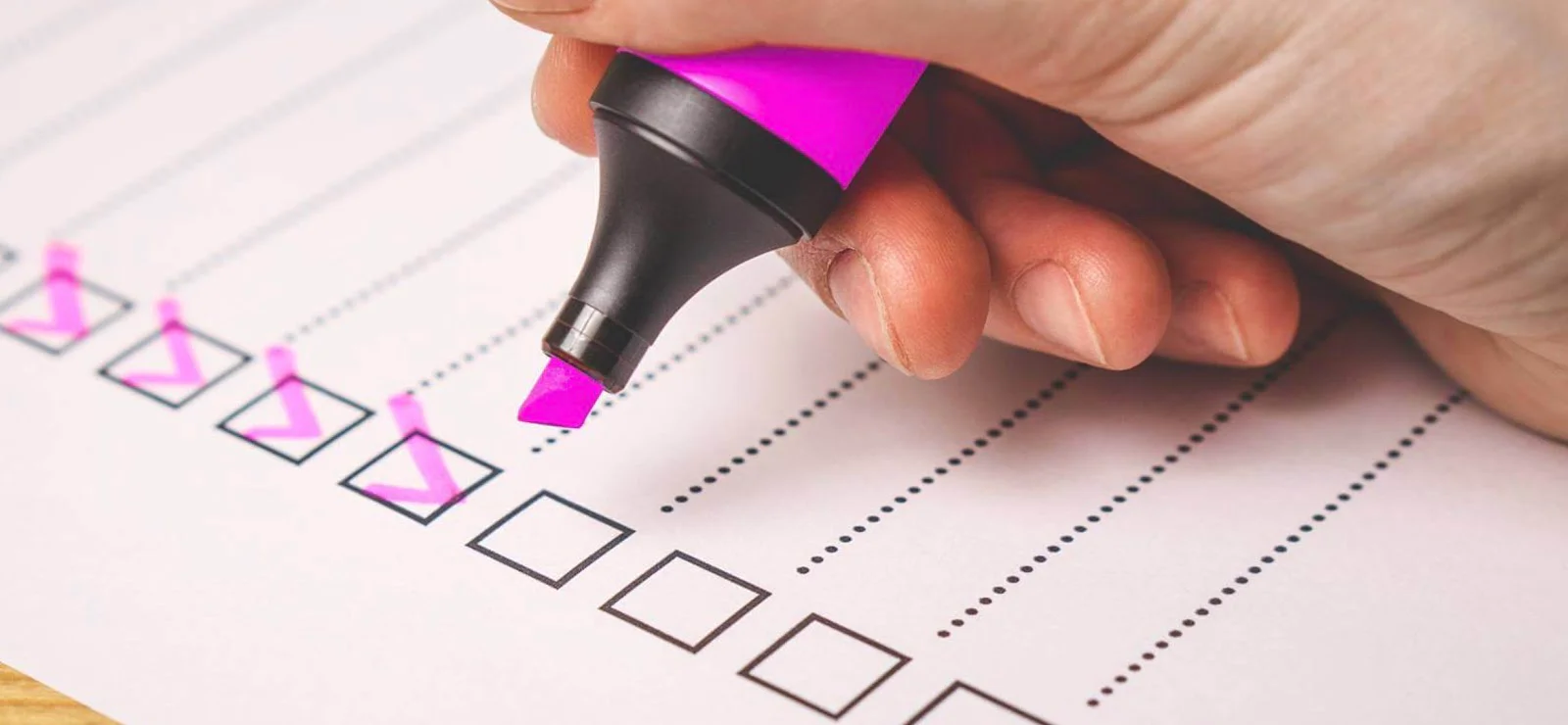 Hand using a pink highlighter to mark checkboxes on a paper checklist.