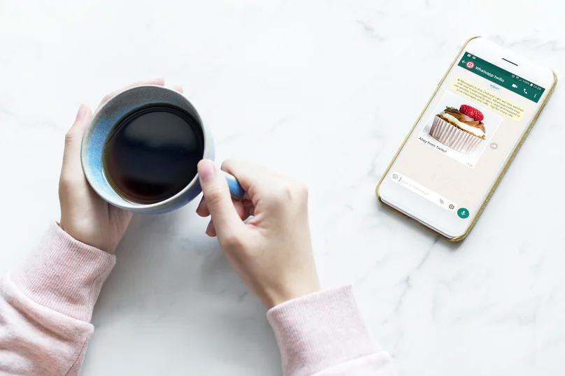 Person holding a cup of coffee next to a smartphone displaying a WhatsApp message with a cupcake image.