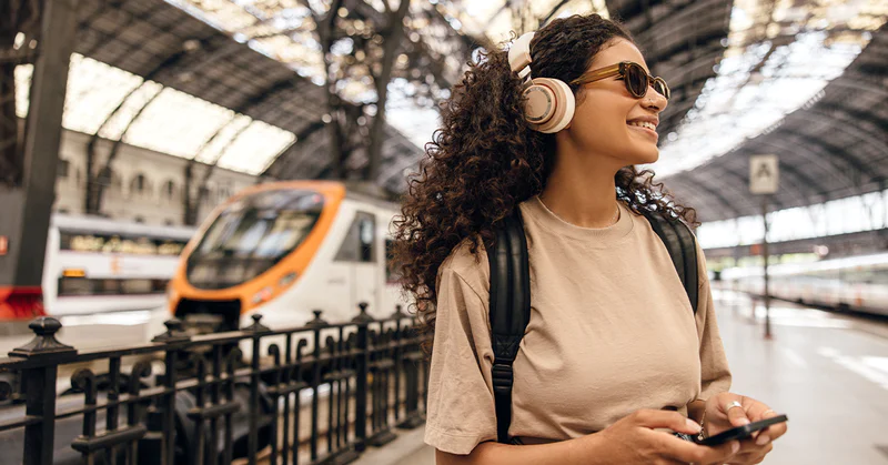 Smiling woman with headphones at a train station holding a phone