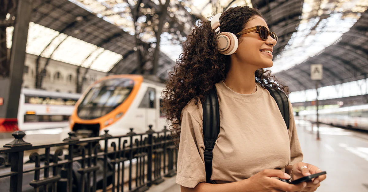 Smiling woman with headphones at a train station holding a phone