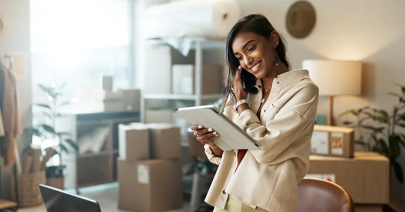 Woman talking on phone and using tablet in a cozy home office with boxes and plants.