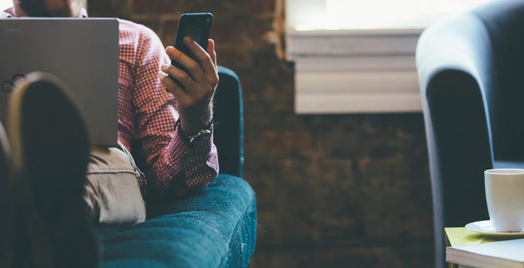 Person sitting on couch with laptop, looking at smartphone in hand, with coffee mug on table.