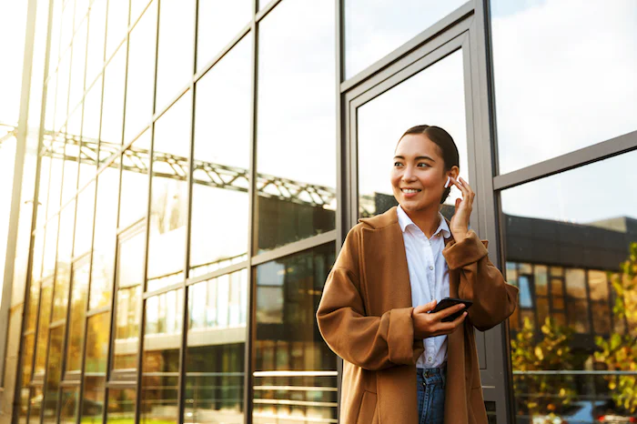Woman in brown coat smiling while using earbuds and holding smartphone outside glass building
