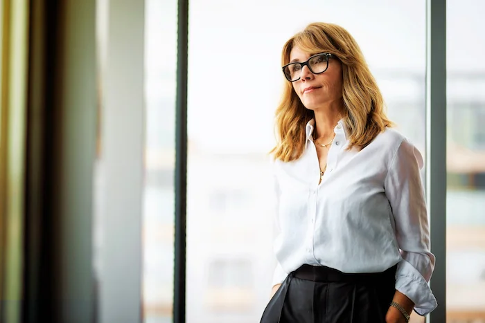 A woman in glasses and a white blouse stands by a window, gazing outside thoughtfully.
