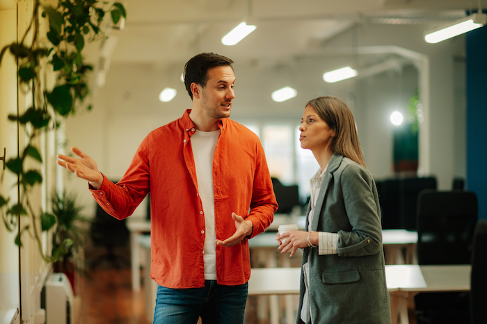 Two colleagues having a conversation in a modern office with plants and computer desks.