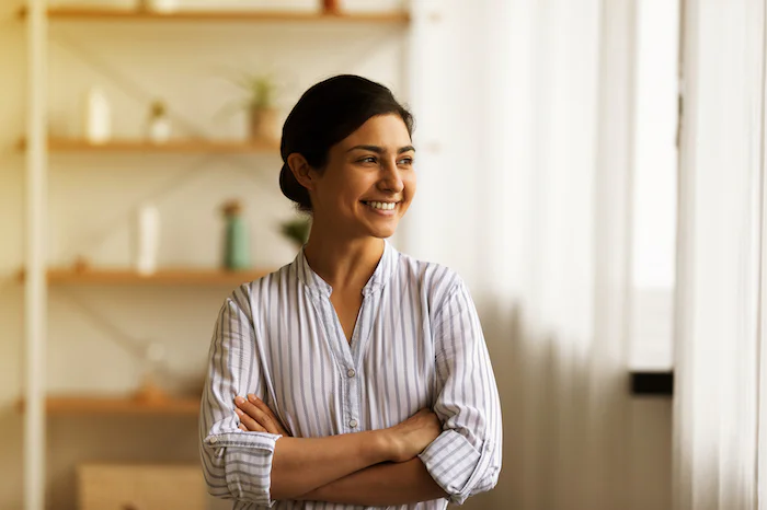 Smiling woman with folded arms looking out a window in a bright room with shelves and plants