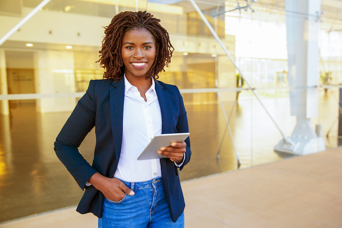 Smiling woman in blazer holding tablet, standing in a brightly lit, modern office building.