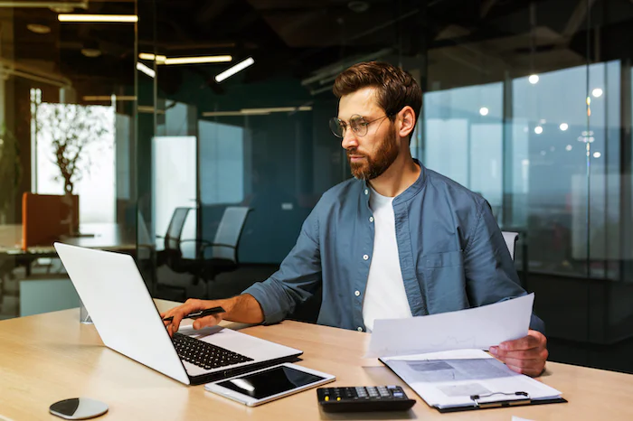 Man working on a laptop while holding documents in an office setting.